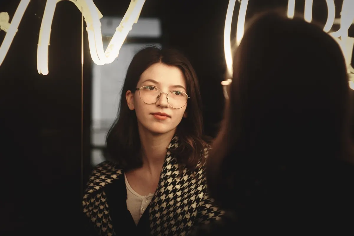 Young woman with glasses looking at her reflection in a lit mirror, thoughtful expression, warm lighting.