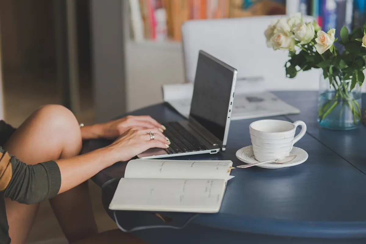 Person typing on a laptop at a table with an open notebook and a cup of coffee, suggesting an online test workspace.