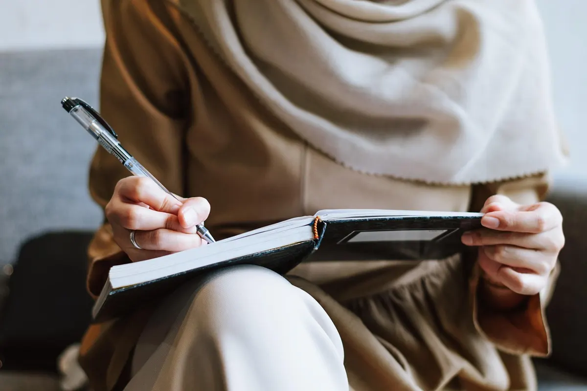 Close-up of hands writing in a notebook on a person's lap with a pen, suggesting reflection and note-taking.