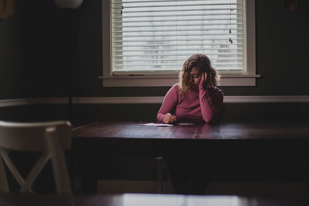 Person sitting alone at a table writing on a paper in a dim room near a window.