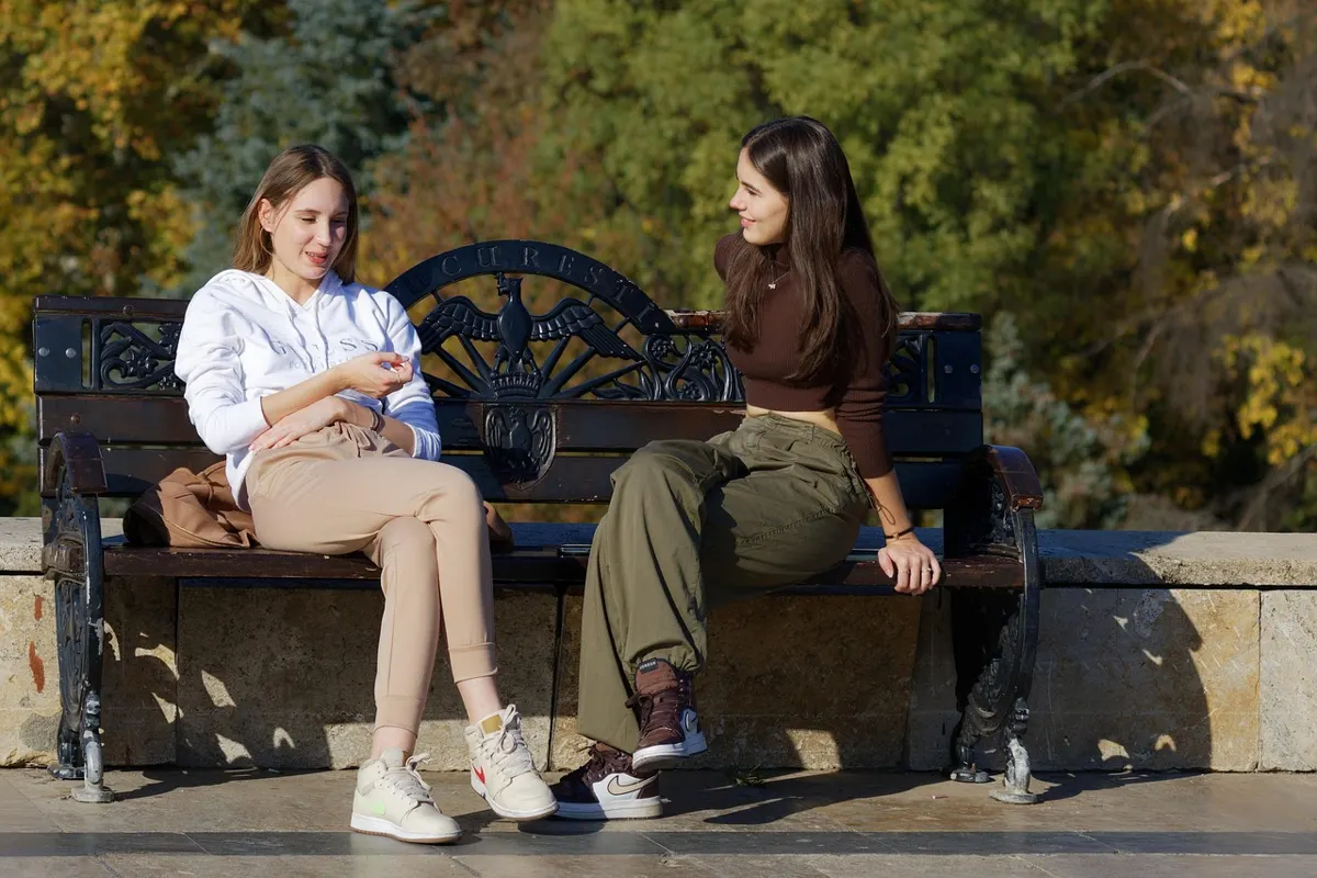 Two young women sitting on a bench outdoors talking and smiling with autumn trees in the background.